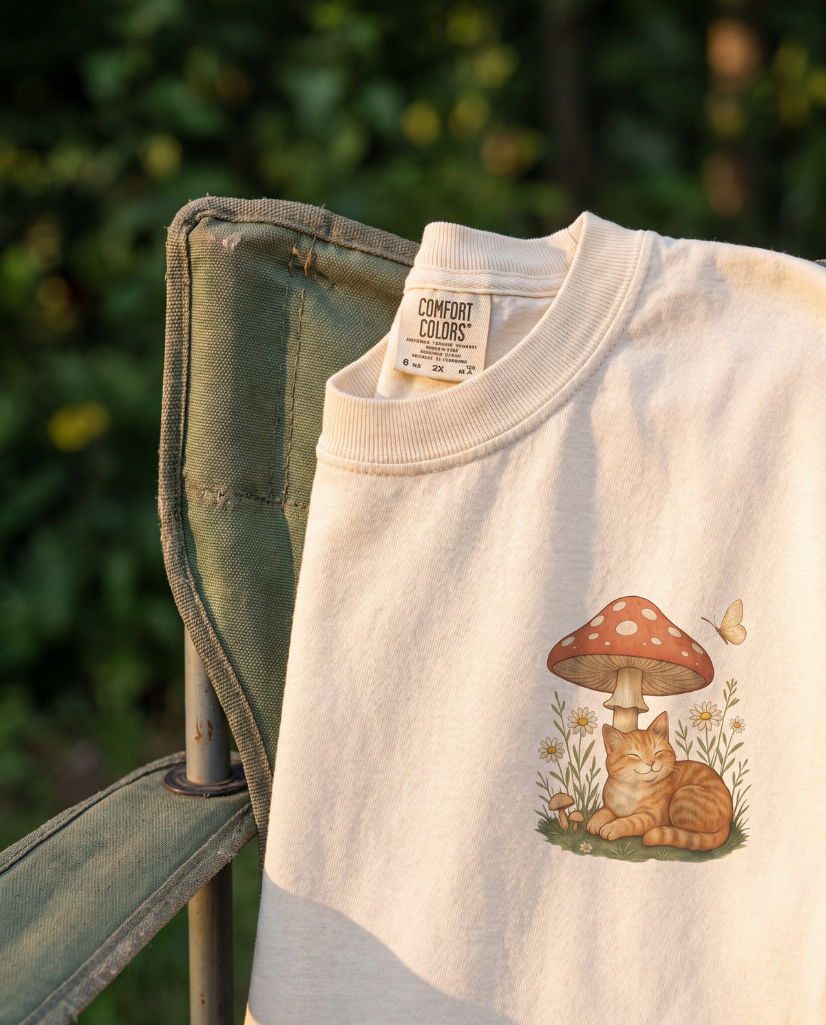 Close-up of an ivory Comfort Colors tee with a sleeping orange cat under a mushroom, draped over a green camp chair at golden hour